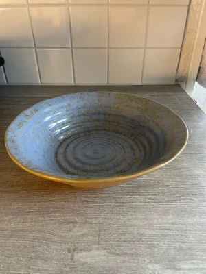 Blå keramikskål med spiral - Säljer en snygg blå keramikskål med spiralformad insida och gulbruna detaljer. Skålen har en rund form och ett unikt mönster som ger den en cool och modern känsla. Perfekt till snacks, sallad eller som dekoration.