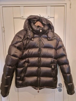 Svart dunjacka från Moncler - Säljer en svart dunjacka från Moncler med glansig finish och vadderad design. Jackan har två dragkedjeförsedda fickor framtill, hög krage och avtagbar huva. Perfekt för kalla vinterdagar och riktigt snygg till streetwear-stilen. Ingen förpackning men pris lapp.