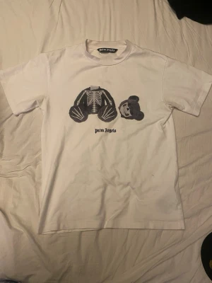Vit Palm Angels t-shirt med björntryck - Säljer en vit t-shirt från Palm Angels med ett ikoniskt tryck av en nallebjörn med skelettdetaljer på bröstet. T-shirten har rund hals och korta ärmar. Perfekt för dig som gillar streetwear och vill sticka ut med en unik design.