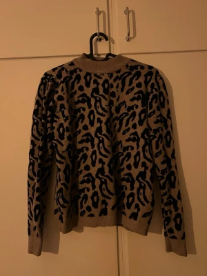 Beige leopardmönstrad stickad tröja - Stickad tröja i beige med svart leopardmönster. Tröjan har rund halsringning, långa ärmar och ribbade muddar vid ärmslut och nederkant. Perfekt för dig som vill ha en trendig och djurinspirerad look.