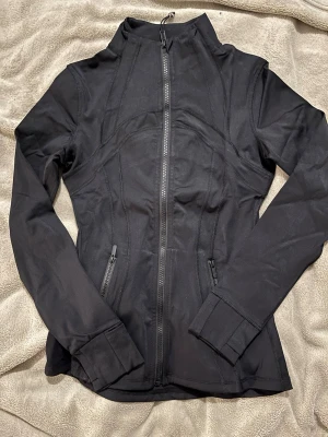 Lululemon define jacket svart - Snygg svart jacka med figurnära passform och hel dragkedja framtill. Jackan har två fickor med dragkedja och hög krage. Materialet är stretchigt och mjukt, perfekt för en aktiv stil. Ärmsluten har breda muddar för extra komfort.