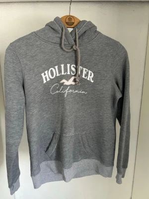 Grå Hollister hoodie med tryck - Snygg grå hoodie från Hollister med vit logga och texten 'California' broderad på bröstet. Klassisk känguruficka framtill, ribbade muddar och justerbar huva med dragsko. Perfekt för en avslappnad och trendig look.