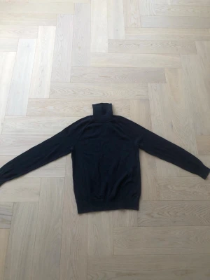 Svart polotröja från Bläck - Klassisk svart stickad polotröja från Bläck med lång ärm och ribbade muddar. Polokragen är hög och mjuk, perfekt för lager på lager. Enkel och stilren design som passar till det mesta. Materialet känns mjukt och följsamt.