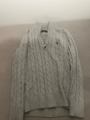 Grå stickad tröja Polo Ralph Lauren - Stickad grå tröja från Polo Ralph Lauren med halv dragkedja och ribbade muddar. Klassisk logga broderad på bröstet. Tröjan har hög krage och är gjord i mjukt material som passar perfekt till kyliga dagar. Pris kan även diskuteras så skriv jättejärna vid eventuella frågor!