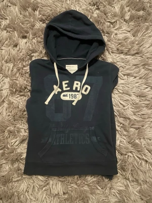 Mörkblå Aeropostale hoodie med tryck - Mörkblå hoodie från Aeropostale med stor huva och vita dragsnören. Framtill finns ett stort tryck med texten 'AERO 87' och 'ATHLETICS'. Hoodien har en klassisk känguruficka och är gjord i mjukt bomullsmaterial. Perfekt för en avslappnad streetstyle.