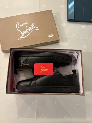 Svarta Louboutin sneakers i läder - Snygga svarta sneakers från Christian Louboutin i slätt läder. Klassisk låg modell med diskreta detaljer och svart sula. Insidan är beige och skorna har en stilren look som passar till det mesta. Levereras i originalkartong.