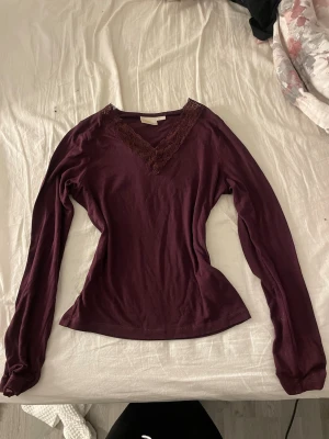 Vinröd långärmad topp Gina Tricot - Vinröd långärmad topp från Gina Tricot med v-ringning och spetsdetaljer runt halsen. Tillverkad i mjukt material som känns skönt mot huden. Perfekt för dig som gillar en enkel men ändå snygg look.