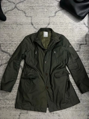 Olivgrön vindjacka från J.Lindeberg - Snygg olivgrön vindjacka från J.Lindeberg med två bröstfickor med dragkedja och två stora fickor med lock. Jackan har klassisk krage, diskret logotyp på ärmen och stängs med både dragkedja och knappar. Materialet är glansigt och vattenavvisande.