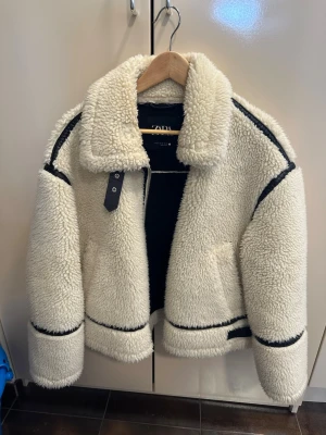 Vit teddyjacka från Zara - Säljer en fluffig och trendig vit teddyjacka från Zara med svarta detaljer längs sömmarna. Jackan har stor krage, två fickor och markerade muddar. Perfekt för dig som vill ha en mysig och snygg jacka till hösten eller vintern.