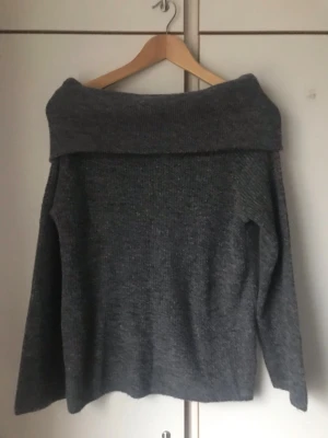 Mörkgrå off shoulder stickad tröja - Säljer en mörkgrå stickad tröja med bred off shoulder-krage. Tröjan har lång ärm och är tillverkad i mjukt, tjockt material som passar perfekt för kyligare dagar. Enkel och stilren design utan synligt märke.