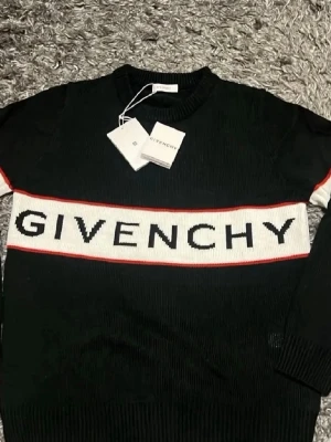 Svart stickad tröja från Givenchy - Svart stickad tröja från Givenchy med vit panel och stor logotyp över bröstet. Tröjan har röda detaljer längs panelen, rund halsringning och ribbade muddar. Perfekt för dig som gillar exklusiv streetwear och vill sticka ut.