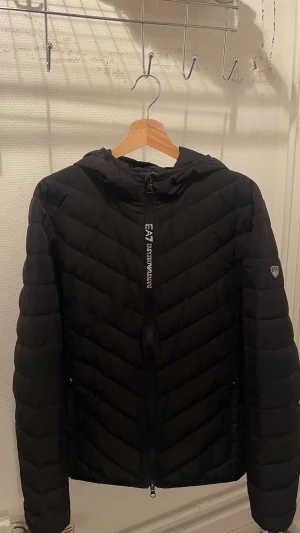 Svart pufferjacka från EA7 Emporio Armani - Snygg svart pufferjacka från EA7 Emporio Armani med huva och dragkedja framtill. Jackan har ett stilrent märke på ärmen och diskret logga längs dragkedjan. Perfekt för kalla dagar och ger en sportig vibe. 