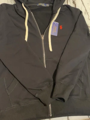 Ralph lauren hoodie  - Snygg svart hoodie från Polo Ralph Lauren med dragkedja, vit dragsko och klassisk röd logga på bröstet. Tillverkad i mjuk bomull och har två fickor framtill samt ribbade muddar vid ärmslut och nederkant. Perfekt för en chill och stilren look.