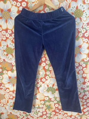 Blå sammetsmjukisbyxor Juicy Couture - Mörkblå mjukisbyxor i mjuk sammet från Juicy Couture. Byxorna har en bred resår i midjan och raka ben. Liten JC-logga broderad vid linningen. Perfekta för en chill och avslappnad stil.