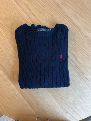 Mörkblå kabelstickad tröja Polo Ralph Lauren - Kabelstickad mörkblå tröja från Polo Ralph Lauren med rund halsringning och röd broderad logga på bröstet. Tröjan har ribbade muddar vid ärmslut och nederkant. Klassisk och tidlös design i mjukt material.