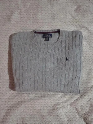 Grå kabelstickad tröja Polo Ralph Lauren - Kabelstickad grå tröja från Polo Ralph Lauren med rund halsringning och klassisk broderad logga på bröstet. Tröjan har långa ärmar och ribbade muddar vid ärmslut och nederkant. Perfekt för dig som gillar stilrena och tidlösa plagg. Längd 60cm & bredd 40 cm