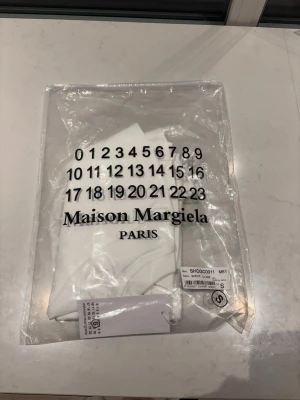 Vit Maison Margiela t-shirt med tryck - Säljer en vit Maison Margiela t-shirt från med det ikoniska siffertrycket och loggan på bröstet. T-shirten har rund hals, korta ärmar och är gjord i mjuk bomull. Perfekt för dig som gillar minimalistisk och stilren design.
