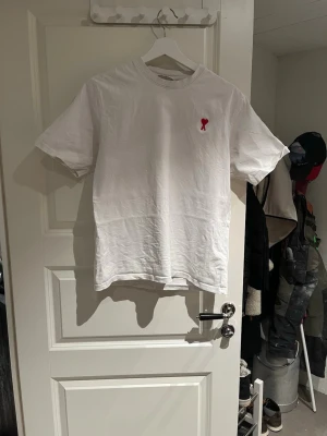Vit t-shirt från Ami Paris  - Säljer en klassisk vit t-shirt från Ami Paris med röd broderad hjärtdetalj och bokstaven A på bröstet. T-shirten har rund hals och korta ärmar, perfekt till alla dina outfits. Tillverkad i mjuk bomull för skön känsla. Samt att den har ett litet håll men använder själv den med hållet och ingen lägger märke till det! 