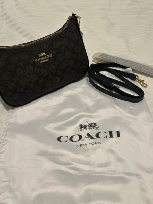 Coach väska - Perfekt Skick! - Snygg väsk från Coach med klassiskt monogrammönster och guldfärgade detaljer. Väskan har avtagbar och justerbar axelrem i svart läder samt dragkedja upptill. Perfekt storlek för det viktigaste och levereras med dustbag.