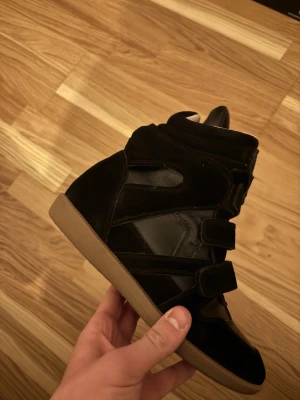 Svarta isabel marant - Svarta stövletter med högt skaft, flera breda kardborreband och detaljer i mocka och slätt material. Skorna har beige gummisula och vadderad insida för extra komfort. Perfekt för dig som gillar en sportig och edgy look.