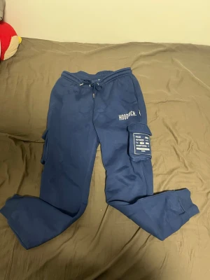 Blå cargo mjukisbyxor från Hoodrich - Säljer ett par blå mjukisbyxor från Hoodrich med cargo-fickor på sidorna och dragsko i midjan. Byxorna har tryckt logga på vänster ben och en patch med text på fickan. Sköna och stilrena, perfekta för chill dagar.