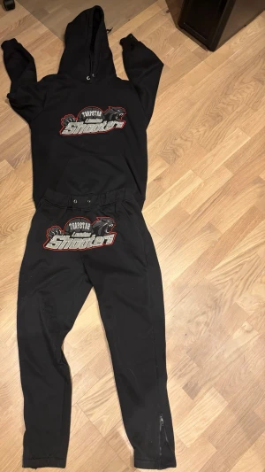 Trapstar London Shooters svart set - Svart loungewear-set från Trapstar med tryck 'London Shooters' och pantermotiv på både hoodie och byxor. Hoodien har dragsko och byxorna har resår i midjan samt dragkedja vid benslut. Perfekt för chill dagar hemma.