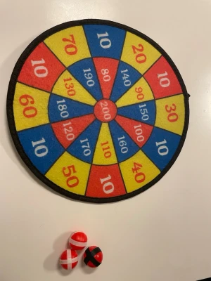 Färgglad darttavla i tyg med bollar - Säljer en rund darttavla i tyg med färgglada fält i rött, gult och blått samt poäng i varje sektion. Tavlan kommer med tre mjuka bollar i rött och svart som fäster på tavlan. Perfekt för spel hemma eller på festen!
