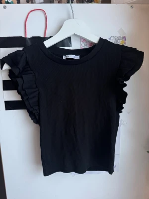 Svart volangtopp från Zara - Säljer en svart ribbad topp från Zara med volangdetaljer på ärmarna. Toppen har rund halsringning och är tillverkad i ett mjukt, stretchigt material som sitter snyggt på kroppen. Perfekt för dig som vill ha en enkel men ändå trendig look.