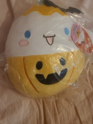 Squishmallows Cinnamoroll pumpa - Mjukisdjur från Squishmallows föreställande Cinnamoroll utklädd till pumpa med Halloween-tema. Färgerna är vit, gul och svart med broderade detaljer och en glad pumpa på magen. Märkt med Hello Kitty and Friends-tag.