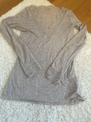 Beige loungewear-topp med spets - Mjuk och tunn beige loungewear-topp från Intimissimi med lång ärm, v-ringning och snygga spetsdetaljer vid halsringning och ärmslut. Perfekt för chill dagar hemma eller som ett extra lager under en hoodie. 