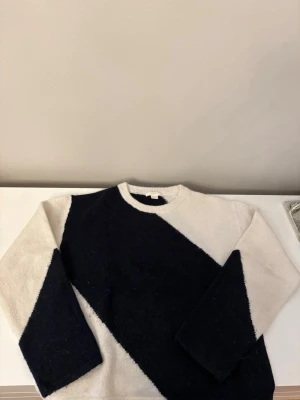 Svartvit stickad tröja  - Säljer en stickad tröja i svart och vitt med en cool diagonal färgblock-design. Tröjan har rund halsringning och långa ärmar. Perfekt för dig som gillar en stilren men ändå unik look.