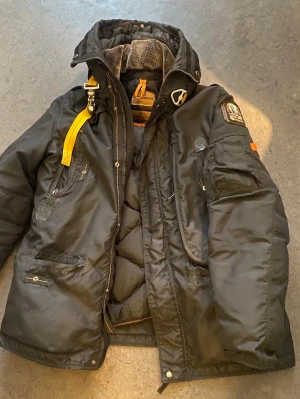 Svart dunjacka från Parajumpers - Säljer en svart dunjacka från Parajumpers med ikonisk gul rem vid kragen, flera fickor och patch på ärmen. Jackan har hög krage med mjukt foder och robust dragkedja. Perfekt för kalla vinterdagar och riktigt snygg streetstil.