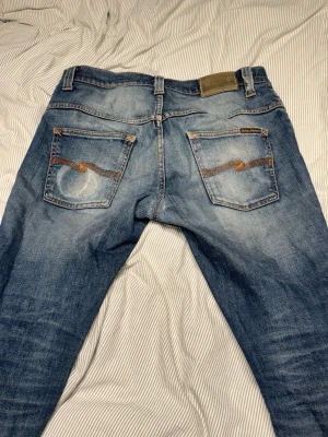 Blå jeans från Nudie Jeans - Säljer ett par klassiska blå jeans från Nudie Jeans med snygg slitning och kontrastsömmar. Jeansen har fem fickor, orange broderi på bakfickorna och en patch i midjan. Perfekta för en avslappnad och trendig look.