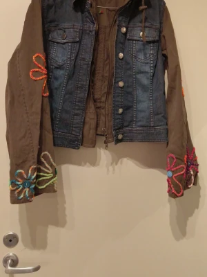 Unik jeansjacka med broderade blommor - Säljer en cool jeansjacka med bruna tygärmar och huva. Jackan har färgglada broderade blommor och mönster på ärmar, huva och rygg. Dragkedja och knappar framtill, två bröstfickor. Perfekt för dig som vill sticka ut med din stil! Storlek 38-40