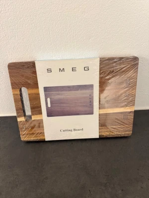 Smeg skärbräda i trä oanvänd och ej öppnad  - Snygg skärbräda från Smeg i mörkt trä med ljusare trädetalj och ingraverad logga. Rektangulär form med praktiskt handtag på sidan. Perfekt för köket när du vill ha både stil och funktion. Måtten är 32x23x1.7