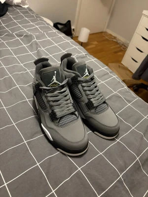Gråa Air Jordan 4 sneakers - Snygga Air Jordan 4 sneakers i olika grå nyanser med vita och svarta detaljer. Skorna har meshpaneler, klassisk snörning och Jumpman-logga på plösen och hälen. Tillverkade i en mix av läder och syntetmaterial för en clean och sportig look.