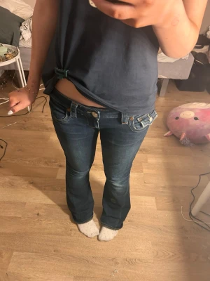 Joey true religion W27 - Säljer ett par mörkblå bootcut jeans med snygga kontrastsömmar och dekorativa ficklock bak. Jeansen har låg midja och klassisk femficksdesign. Perfekta för dig som gillar en retro vibe och vill ha något som sticker ut lite extra.