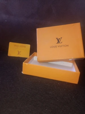 Louis Vuitton ask och kort - Säljer en orange ask från Louis Vuitton med tillhörande kort. Asken har klassisk LV-logga på locket och är gjord i hård kartong. Perfekt för förvaring av småsaker eller som presentförpackning.