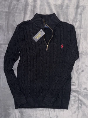 Svart kabelstickad tröja Polo Ralph Lauren - Kabelstickad svart tröja från Polo Ralph Lauren med halv dragkedja och hög krage. Tröjan har det klassiska röda Polo-logot broderat på bröstet och ribbade muddar vid ärmslut och nederkant. Perfekt för lager på lager och snygg till jeans.