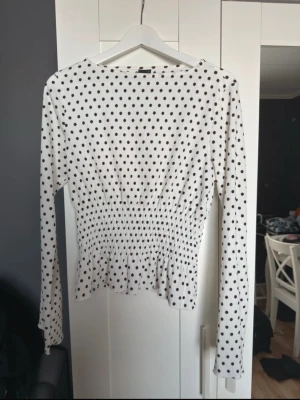 Vit prickig blus  - Snygg vit blus med svarta prickar och smockad midja som ger en figurnära siluett. Blusen har långa ärmar och en lätt volang nedtill. Perfekt för dig som gillar trendiga och feminina detaljer. Ny utan prislapp!