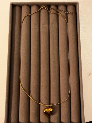 Guldigt halsband med hjärthänge - Snyggt halsband i guldfärgad metall med ett blankt, tredimensionellt hjärthänge. Kedjan är tunn och elegant med justerbar längd. Perfekt accessoar för att lyfta vilken outfit som helst. Hjärtat ger en romantisk touch och halsbandet känns både trendigt och stilrent.
