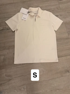 Vit ribbad t-shirt med half zip - Säljer en stilren vit t-shirt från Weekday i ribbat material. T-shirten har korta ärmar, krage och en snygg half zip-dragkedja framtill. Perfekt för dig som gillar minimalistisk och modern stil.
