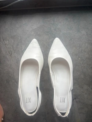 Vita ballerinaskor med öppen häl - Säljer ett par stilrena vita ballerinaskor från XIT med spetsig tå och öppen häl. Skorna har låg klack och en minimalistisk design som passar till många olika outfits. Tillverkade i ett slätt material som ger en clean look.