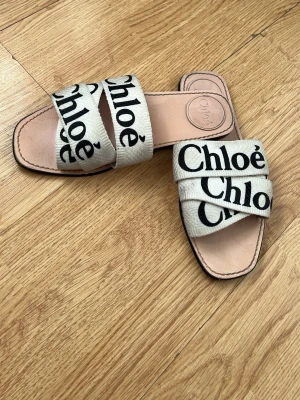 Beige sandaler från Chloé - Snygga beige sandaler från Chloé med två breda tygband över foten, dekorerade med svart logotyptext. Sandalerna har fyrkantig tå och innersula i ljust läder. Perfekta för en trendig och avslappnad look. Detta är en high copy och inte original. Originalet kostar 4700kr