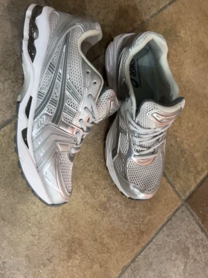 Silvriga Asics sneakers - Snygga sneakers från Asics i silver och vitt med mesh- och syntetmaterial. Skorna har en sportig design med tydliga Asics-loggor på sidorna och en bekväm, vadderad insida. Perfekta för dig som gillar retrovibbar och streetstyle.