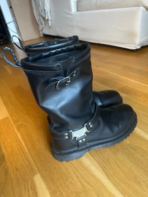 Svarta läderboots från Dr. Martens - Tuffa svarta boots i läder från Dr. Martens med grov sula, metallspänne och dekorativa remmar. Klassisk hög modell med dragflikar bak och robust känsla. Perfekta för dig som vill ha en edgy stil.