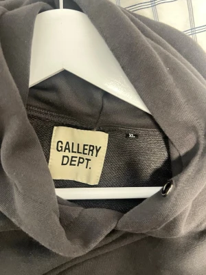 Mörkgrå hoodie från Gallery Dept. - Säljer en mörkgrå hoodie från Gallery Dept. i storlek XL. Hoodien har en klassisk huva och ribbade muddar. På ärmen sitter en vit patch med texten 'ART ON DISPLAY Courtesy of JOSUÉ THOMAS'. Perfekt för dig som gillar streetwear och unik design.