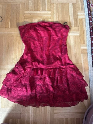 Röd axelbandslös volangklänning - Snygg röd axelbandslös klänning med volangdetaljer och blommigt mönster. Perfekt för dig som vill sticka ut! Klänningen har en kort längd och är tillverkad i ett lätt, något glansigt material som ger en lyxig känsla.