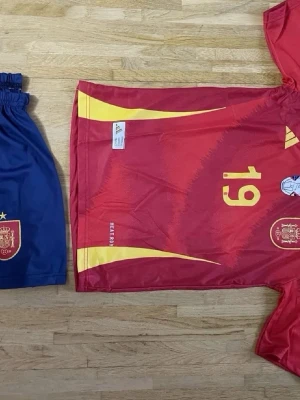Spaniens matchställ Adidas röd/blå - Säljer ett matchställ från Spaniens landslag med röd tröja och gula detaljer, nummer 19 på bröstet och officiella landslagsmärken. Tillhörande blå shorts med guldfärgat landslagsemblem och stjärna. Tillverkat i lätt och ventilerande material.