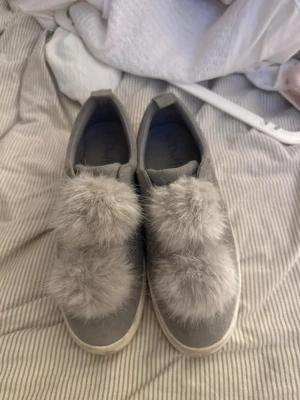 Grå sneakers med fluffiga bollar - Säljer ett par grå sneakers från Duffy med stora fluffiga bollar i fuskpäls på ovansidan. Skorna har en låg modell och vit sula. Materialet är mockaimitation och insidan är mjuk för extra komfort. Perfekta för dig som vill sticka ut med din stil.